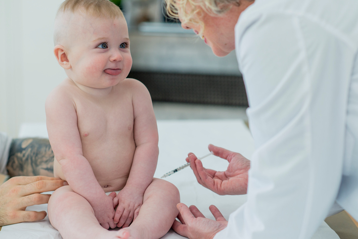 A baby boy sits patiently while a doctor administers a vaccine. The baby boy is at the doctor's office for a checkup. The baby boy smiles up at his doctor.