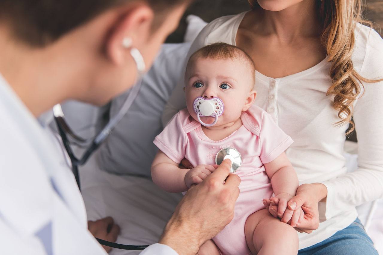 Beautiful young mom is holding her cute baby while doctor is listening to baby's lungs