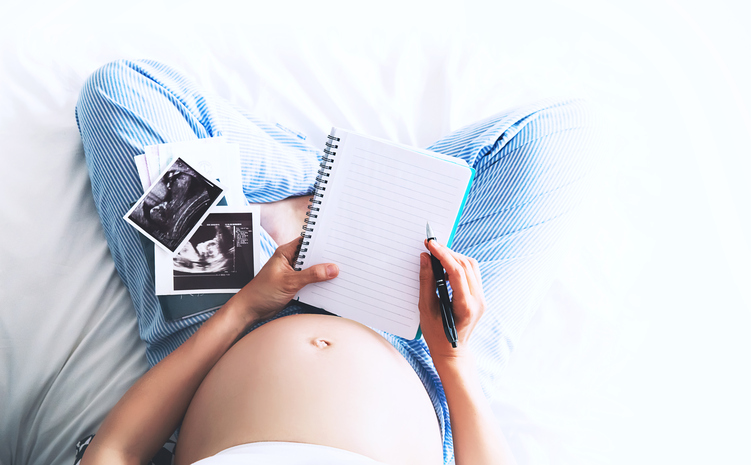 Pregnant woman makes notes in notebook and holding ultrasound image and medical documents at home interiors. Pregnancy, parenthood, preparation and expectation concept. Close-up, copy space, indoors.