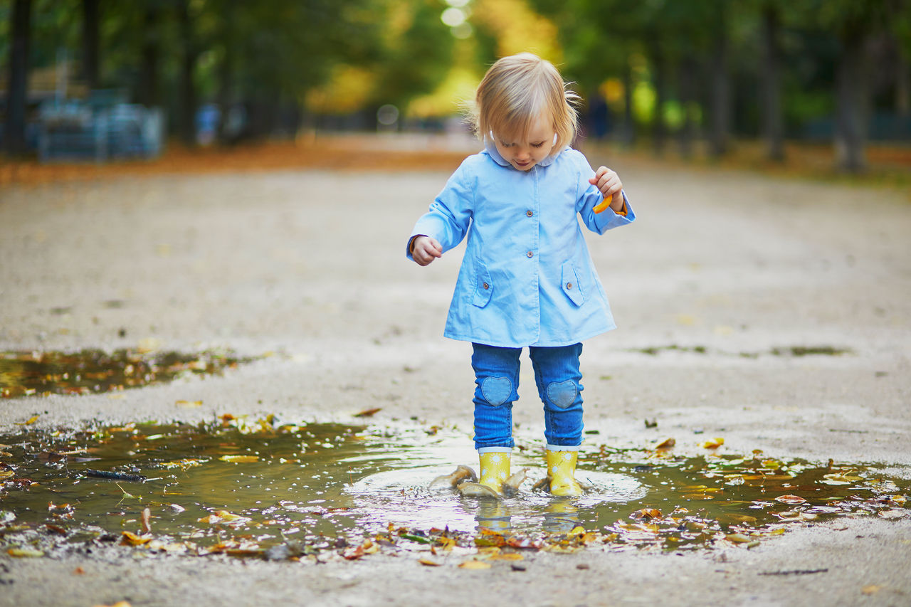 Child wearing yellow rain boots and jumping in puddle on a fall day. Adorable toddler girl having fun with water and mud in park on a rainy day. Outdoor autumn activities for kids